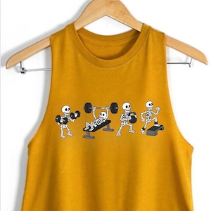 May include: A mustard yellow sleeveless crop top featuring a graphic print of four white skeletons performing different exercises, including lifting dumbbells, bench pressing a barbell, holding a kettlebell, and stepping onto a platform.