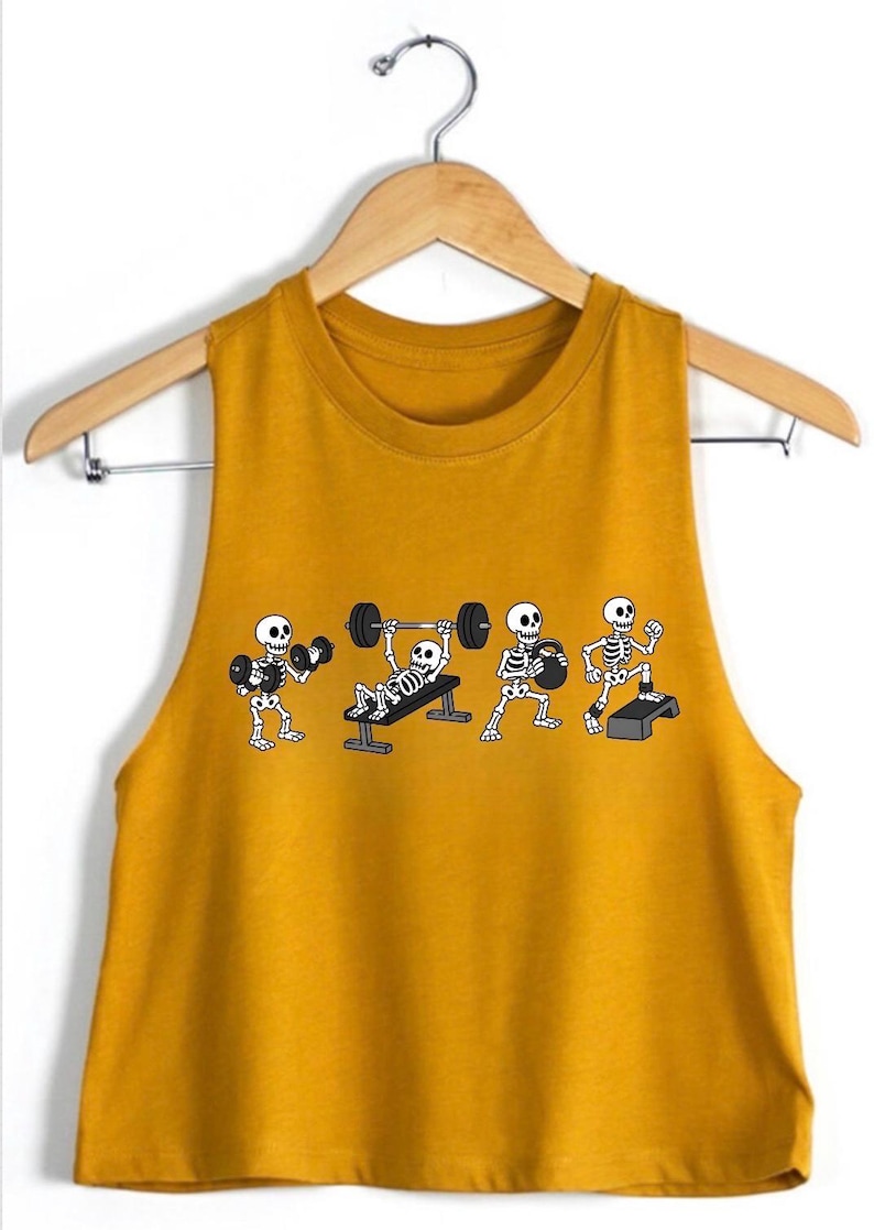 May include: A mustard yellow sleeveless crop top featuring a graphic print of four white skeletons performing different exercises, including lifting dumbbells, bench pressing a barbell, holding a kettlebell, and stepping onto a platform.