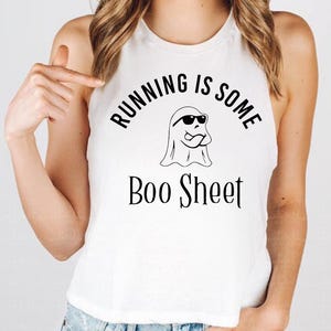 May include: A white sleeveless tank top featuring a black graphic print of a ghost wearing sunglasses. The text above the ghost reads RUNNING IS SOME and the text below the ghost reads Boo Sheet in a stylized black font on the front of the shirt.