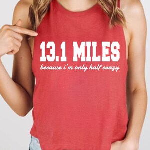 May include: A red sleeveless tank top featuring the white text 13.1 MILES in a bold block font, followed by the white script text because i'm only half crazy. The garment has a round neckline and a cropped hemline, showcasing a simple design.