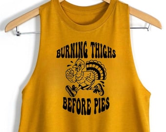 May include: A mustard yellow sleeveless crop top featuring a black graphic of a running turkey wearing trainers. The text above the turkey reads "BURNING THIGHS" and the text below the turkey reads "BEFORE PIES" in a bold, wavy black font.