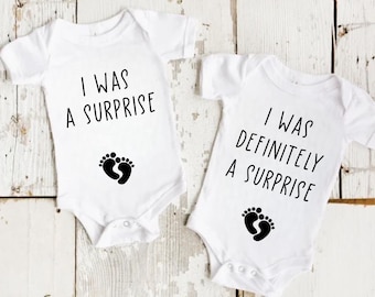 May include: Two white baby onesies with black text and foot prints. One says "I WAS A SURPRISE" and the other says "I WAS DEFINITELY A SURPRISE". The onesies are short-sleeved and have snaps at the bottom.