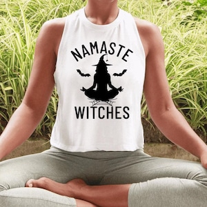 May include: A white sleeveless tank top featuring a black graphic print of a witch in a lotus yoga pose. The text NAMASTE is arched above the witch, and the word WITCHES is printed below. Two small black bats flank the witch silhouette.