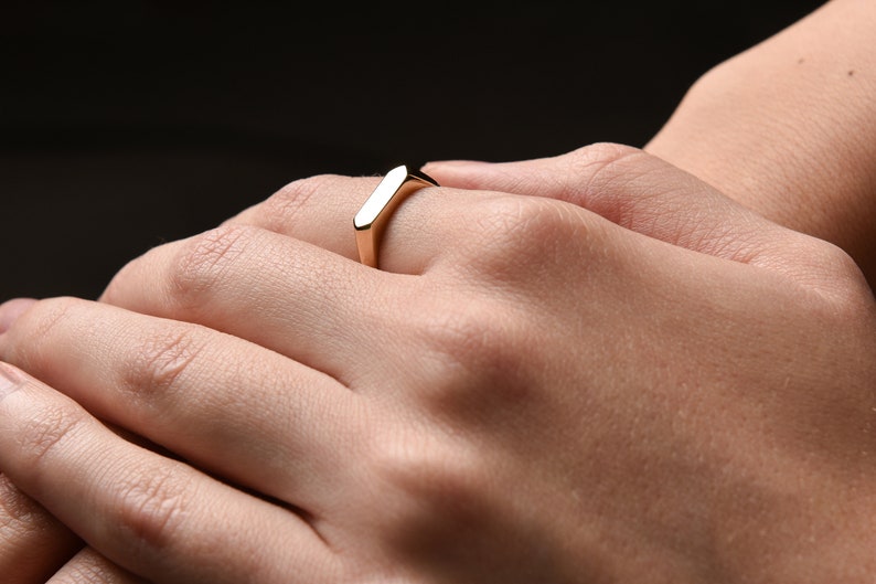 May include: A gold-colored ring with a rectangular band. The ring is worn on a person's finger.