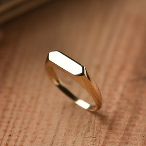 May include: A gold signet ring with a flat, rectangular top. The ring is made of polished metal and has a simple, modern design.