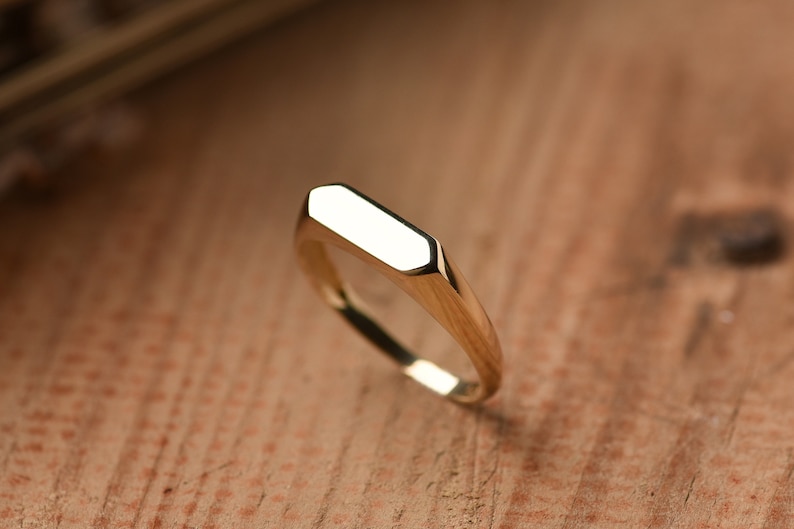 May include: A gold signet ring with a flat, rectangular top. The ring is made of polished metal and has a simple, modern design.
