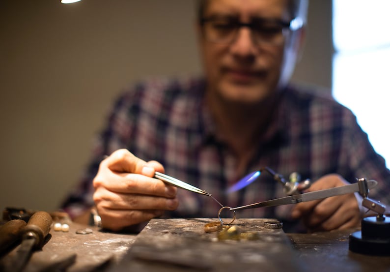 May include: A close-up of a jeweler working on a piece of jewelry with a torch. The jeweler is wearing a plaid shirt and glasses. The jewelry is being held in place with tweezers and is being heated with a torch. The jeweler is working on a workbench with tools and materials.