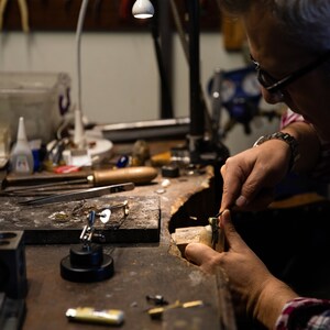 May include: A person working on a piece of jewelry at a workbench. The workbench is covered in tools and materials, including a pair of tweezers, a small hammer, and a piece of wood.