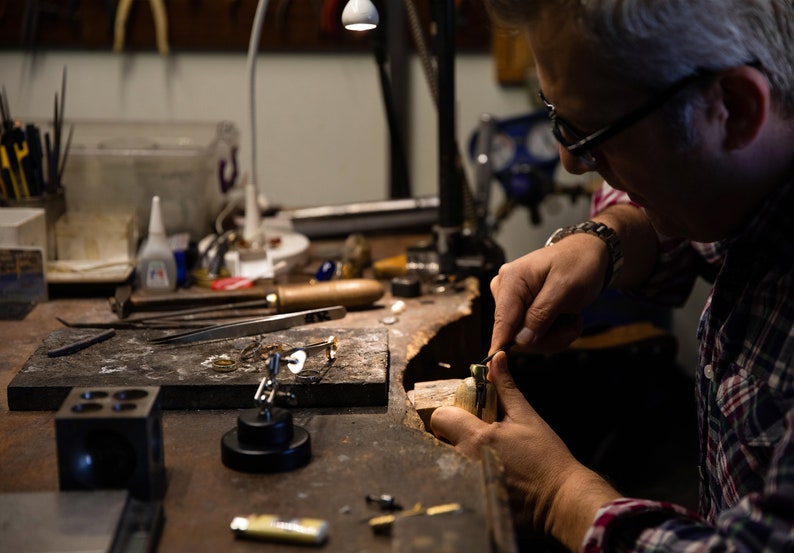 May include: A person working on a piece of jewelry at a workbench. The workbench is covered in tools and materials, including a pair of tweezers, a small hammer, and a piece of wood.