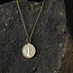 May include: A gold chain necklace with a round pendant featuring a detailed image of a woman holding a staff. The pendant is on a dark gray textured background.
