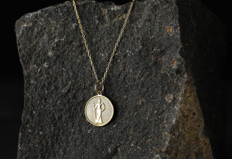 May include: A gold chain necklace with a round pendant featuring a detailed image of a woman holding a staff. The pendant is on a dark gray textured background.