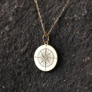 May include: A gold compass necklace with a detailed compass design on a round pendant. The necklace is on a dark gray textured surface.
