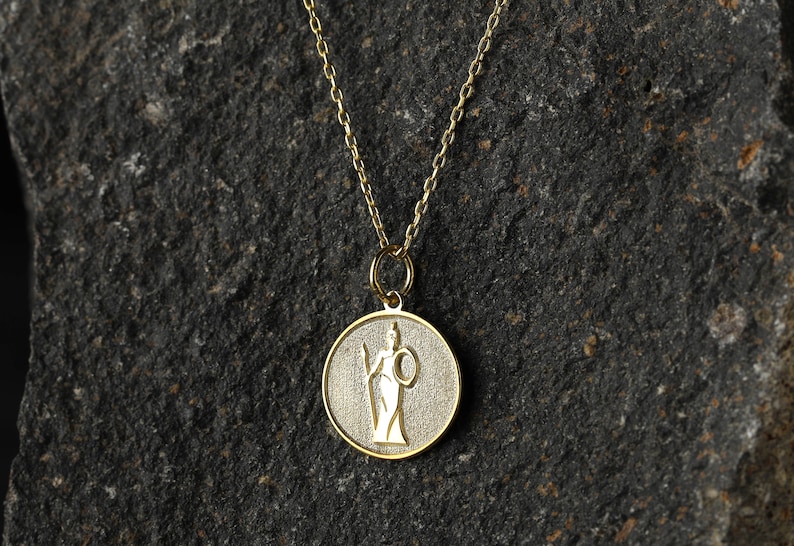 May include: A gold necklace with a round pendant featuring a detailed image of a woman holding a spear and a shield.