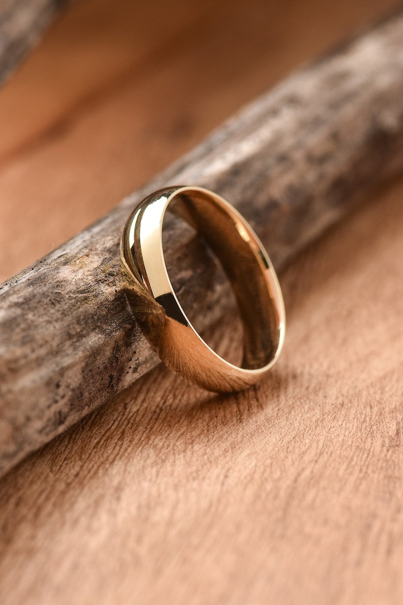 May include: A simple gold wedding band with a polished finish. The band is lying on a piece of wood.