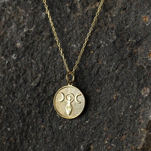 May include: A gold necklace with a round pendant featuring a triple moon symbol and a stylized female figure. The pendant is on a chain against a dark gray background.