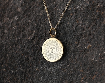 May include: A gold-colored pendant necklace featuring a circular charm with a sun design. The sun has a face in the center and radiating lines. The necklace is on a dark, textured surface.