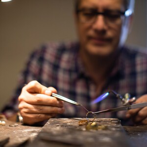 May include: A close-up of a jeweler working on a piece of jewelry with a torch. The jeweler is wearing a plaid shirt and glasses. The jewelry is being held in place with tweezers and is being heated with a torch. The jeweler is working on a workbench with tools and materials.