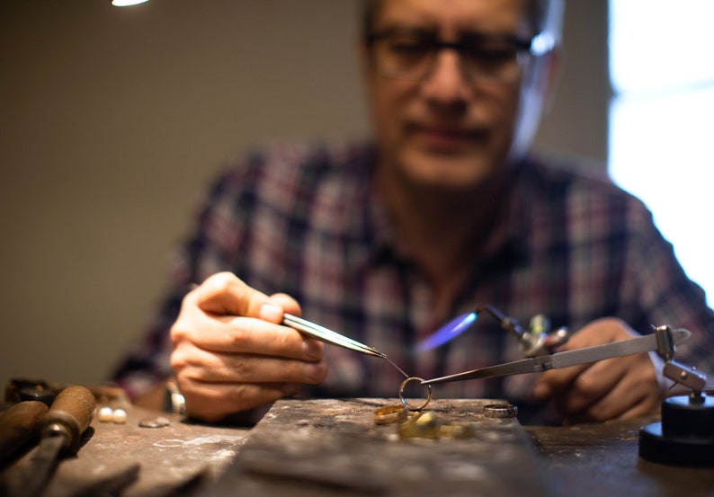 May include: A close-up of a jeweler working on a piece of jewelry with a torch. The jeweler is wearing a plaid shirt and glasses. The jewelry is being held in place with tweezers and is being heated with a torch. The jeweler is working on a workbench with tools and materials.