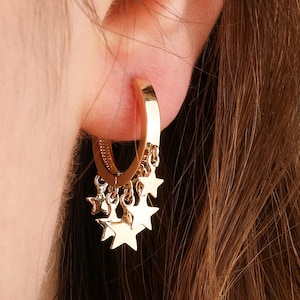 May include: Gold hoop earrings with dangling star charms. The earrings are worn in a person's ear.