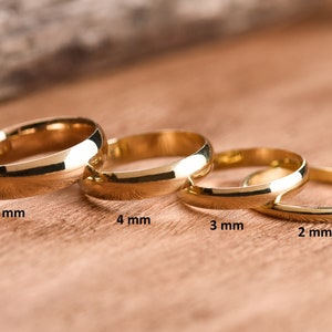May include: Four gold wedding bands of different widths, from 2 mm to 5 mm, laid out on a wooden surface.