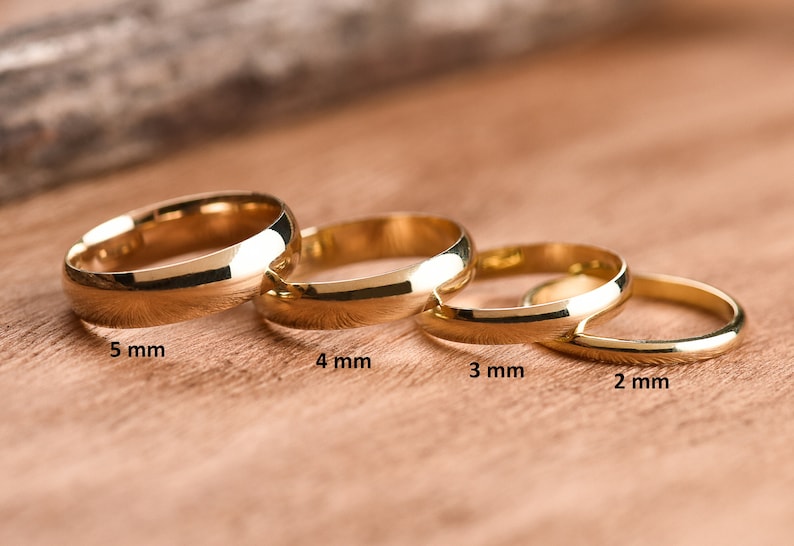 May include: Four gold wedding bands of different widths, from 2 mm to 5 mm, laid out on a wooden surface.
