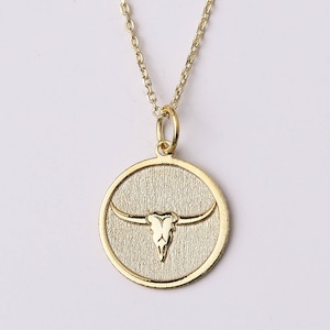 May include: A gold-colored necklace featuring a circular pendant with a textured background and a bull skull design. The pendant is suspended from a delicate chain, creating a simple yet striking piece of jewelry.