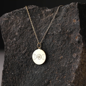 May include: A gold necklace with a round pendant featuring a triple moon and pentagram design. The necklace is displayed on a dark gray rock.