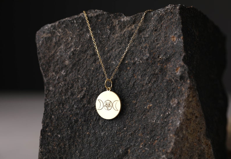 May include: A gold necklace with a round pendant featuring a triple moon and pentagram design. The necklace is displayed on a dark gray rock.