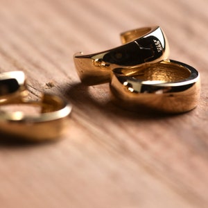 May include: Three gold hoop earrings stacked on top of each other on a wooden surface. The earrings are thick and have a rounded shape.
