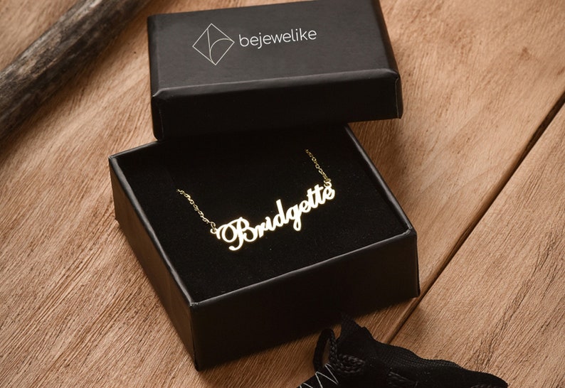 May include: A gold-colored necklace with the name "Bridgette" spelled out in cursive script. The necklace is in a black box with the word "bejewellike" printed on the top.