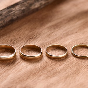 May include: Four gold wedding bands of different sizes are lined up on a wooden surface.
