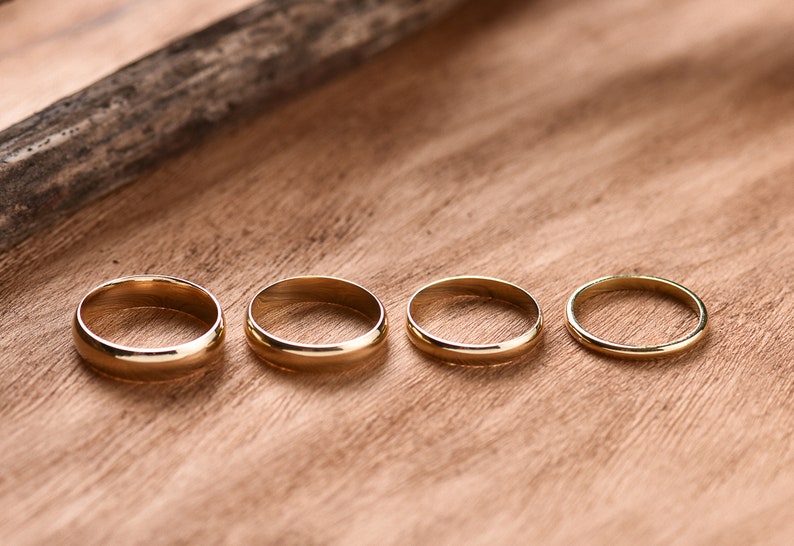 May include: Four gold wedding bands of different sizes are lined up on a wooden surface.