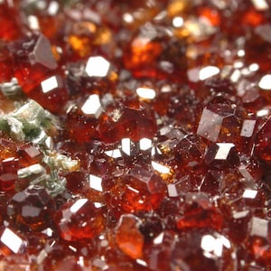 May include: Detailed image of a cluster of deep red garnet crystals. The crystals are faceted and reflect light, with some areas appearing orange-brown. Small patches of green minerals are visible amongst the red crystals.