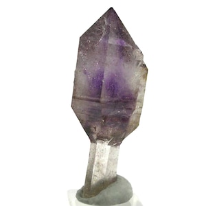 May include: A faceted amethyst crystal with a clear base. The crystal has a deep purple hue in the center, fading to a lighter shade towards the edges. The crystal is displayed on a white background.