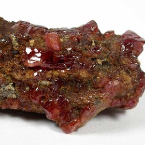 Puede incluir: Un espécimen mineral en bruto que presenta grupos de cristales de color rojo intenso y translúcidos incrustados en una matriz marrón rugosa y texturizada. Las estructuras cristalinas exhiben una superficie brillante y reflectante, que contrasta con el aspecto terroso y granular de la formación rocosa circundante.