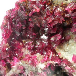 Puede incluir: Primer plano de un espécimen mineral con vibrantes estructuras cristalinas de color rojo intenso. Los cristales están agrupados, creando una superficie texturizada con una mezcla de tonos claros y oscuros. El mineral se encuentra sobre un fondo de material más claro, blanquecino.
