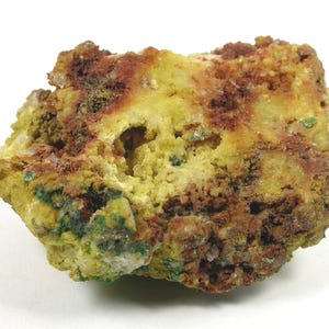 Puede incluir: Un espécimen mineral natural con una textura rugosa y una variedad de colores. La roca presenta una base de amarillo y verde, con áreas de color marrón rojizo y pequeñas formaciones cristalinas. La forma general es irregular.