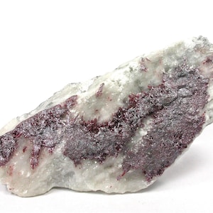 Może przedstawiać: Naturalny okaz minerału z białą matrycą i obszarami ciemnoczerwonych i szarych struktur krystalicznych. Kamień ma szorstki, nieregularny kształt i jest umieszczony na białym tle.