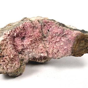 Pode incluir: Uma formação rochosa natural de forma irregular. Apresenta um aglomerado de pequenas estruturas cristalinas rosa. O exterior é uma mistura de castanho, cinzento e verde, contrastando com o interior rosa.