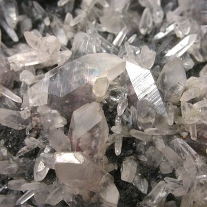 Pode incluir: Um aglomerado de cristais de quartzo translúcidos e incolores com terminações afiadas e pontiagudas e estrias internas visíveis, crescendo densamente sobre uma base de matriz mineral escura e texturizada.