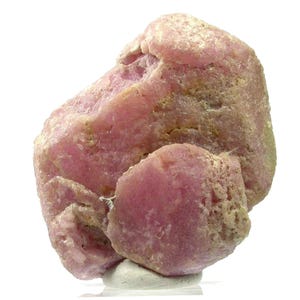 May include: A close-up of a rough, pink mineral specimen. The stone has a textured surface with areas of lighter and darker pink hues, and some areas of tan and beige. The mineral sits on a small, white base against a white background.