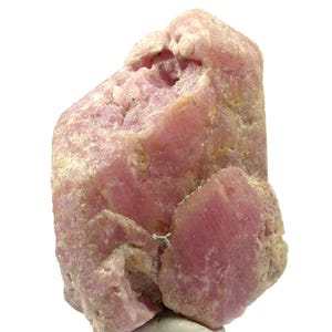 May include: A pink mineral specimen with a rough, crystalline structure. The stone has a light pink hue with areas of darker pink and beige. The mineral is set against a white background.
