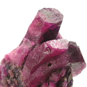 May include: Close-up of a cluster of deep magenta crystals with a rough, textured surface. The crystals have a faceted appearance and are set against a white background. The mineral formation has a natural, organic shape.