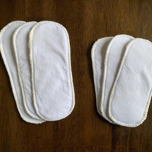 May include: Three white cloth diaper inserts with a white stitched edge. The inserts are stacked on top of each other.