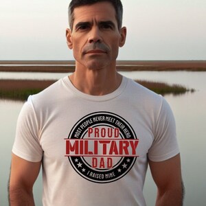 May include: A white t-shirt featuring a black and red circular graphic. The graphic states "Proud Military Dad" with the text "Most people never meet their hero" and "I raised mine". The t-shirt is worn by a person, with a natural background.