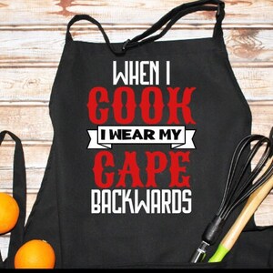 Puede incluir: Delantal negro con texto blanco que dice "When I cook I wear my cape backwards" en letras rojas.