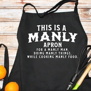 May include: Black apron with white text that reads "This is a Manly Apron for a Manly Man, Doing Manly Things, While Cooking Manly Food."