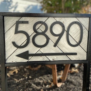 May include: A black metal address sign with the numbers 5890 and an arrow pointing left. The numbers and arrow are mounted on a light gray wooden background with a black frame. The sign is mounted on a black metal stand.
