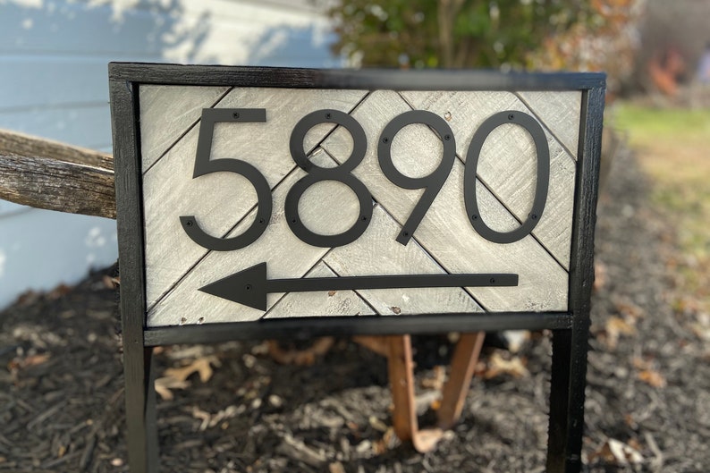 May include: A black metal address sign with the numbers 5890 and an arrow pointing left. The numbers and arrow are mounted on a light gray wooden background with a black frame. The sign is mounted on a black metal stand.