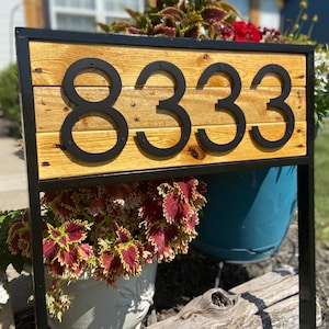 May include: Black metal house number sign with the number 8333 on a wooden plank. The sign is mounted on a black metal frame and is surrounded by plants.